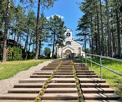 Vertikalna fotografija visokog zvonika Hrama Preobraženja Gospodnjeg, koja vodi pogled na pozlaćeni krst na samom vrhu, dok se u pozadini smjenjuju bijeli oblaci i svijetloplavo planinsko nebo iznad Zlatibora.