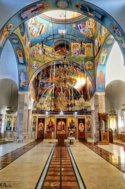 Veličanstveni Hram Preobraženja Gospodnjeg na Zlatiboru, snimljen iz daljine, prikazuje monumentalnu arhitekturu u raškom stilu sa bijelim fasadnim zidovima i prepoznatljivim crvenim krovovima, okružen uređenom promenadom i visokim borovima pod vedrim plavim nebom planinskog turističkog centra.