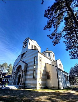 Široki snimak popločanog platoa ispred ulaza u hram na Zlatiboru sa niskim kamenim zidovima i uređenim travnatim površinama, dok se u pozadini uzdiže masivna građevina sa nekoliko manjih kupola i polukružnih apsida.