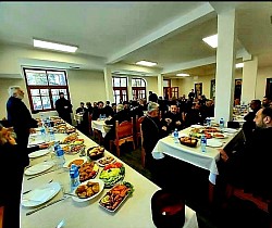 Sabor sveštenstva namesništava užičkog i račanskog za zajedničkom trpezom ljubavi u prostranoj sali parohijskog doma Hrama Preobraženja Gospodnjeg na Zlatiboru nakon bratskog sastanka. Dugi stolovi su postavljeni sa posnom hranom, ribom, salatama i voćem, dok sveštenici u crnim mantijama razgovaraju i zajedničare u svetlom ambijentu