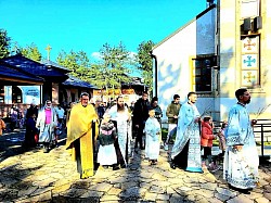 Снимак празничне литије на Златибору поводом Врбице испред цркве Преображења Господњег. У фокусу су велики црквени барјаци од тамноцрвеног сомота са златним везом које носе верници и дечаци у свечаним оделима. На једном од барјака јасно се види вез са иконом Преображења. Литија се креће кроз порту храма, а у позадини се види специфична архитектура златиборске цркве од белог камена са бакарним крововима, уоквирена четинарском шумом и планинским пејзажом. Слика верно преноси саборни дух и очување православне традиције на Краљевском Златибору током априла 2026. године