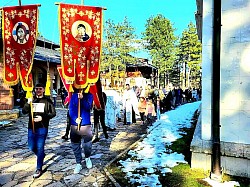 Широк кадар екстеријера Храма Преображења Господњег на Златибору током врхунца прославе Лазареве суботе. Снимљена са висине, фотографија приказује масу људи и деце окупљене у порти храма након литије. Црква, са својом карактеристичном силуетом и крстовима који блистају на сунцу, делује импозантно усред златиборских борова. Видљиви су барјаци и иконе које се носе на челу поворке. Слика савршено документује значај овог верског објекта као духовног центра планине и велику посећеност током православних празника 2026. године.